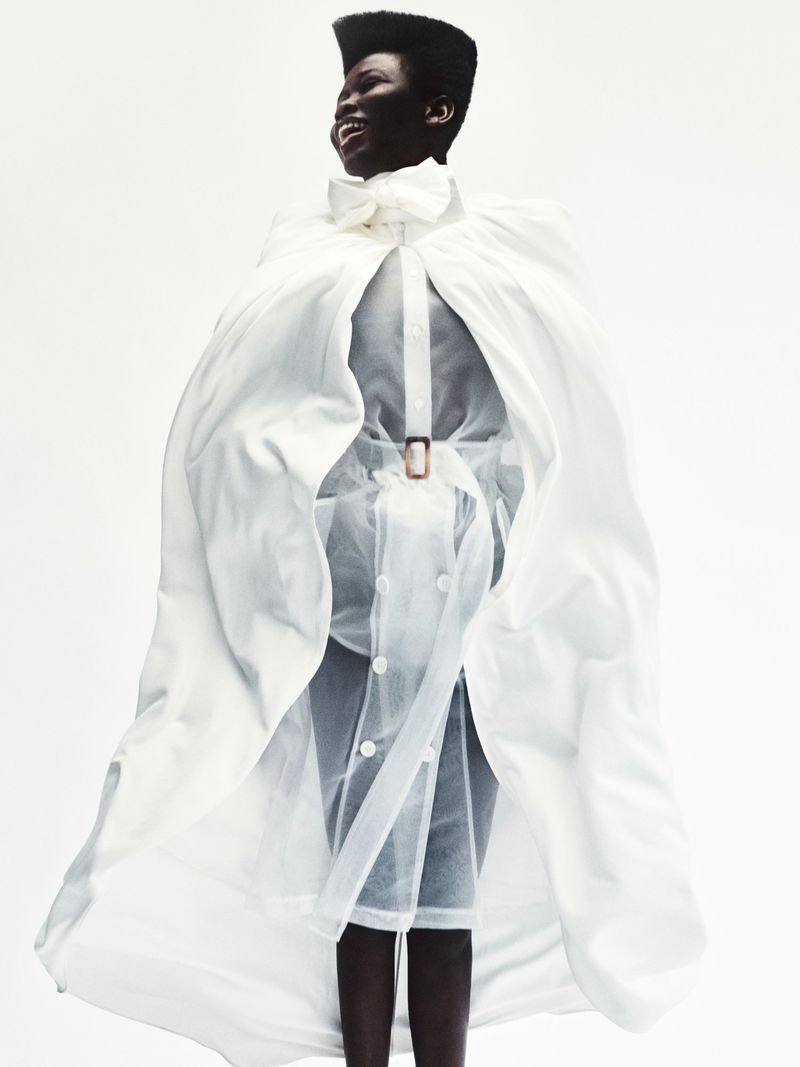 Model Ajah smiling in a dramatic flowing white cape over a sheer buttoned garment against a bright white background.