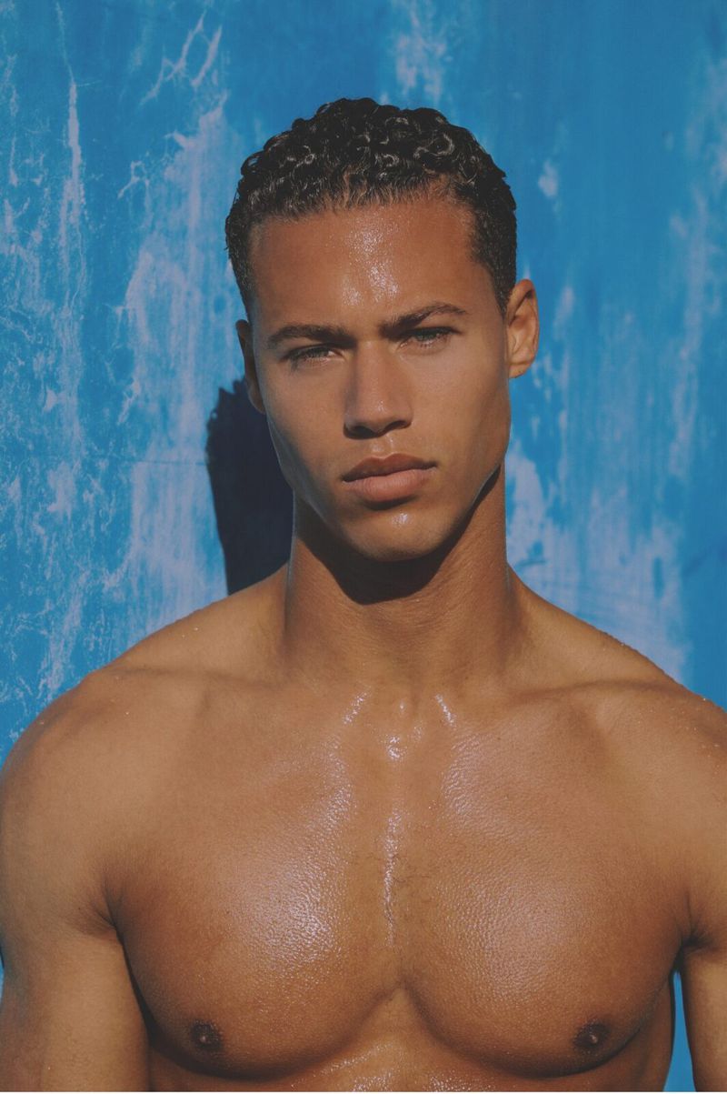Portrait of a shirtless man with short dark curly hair posing against a textured blue wall.
