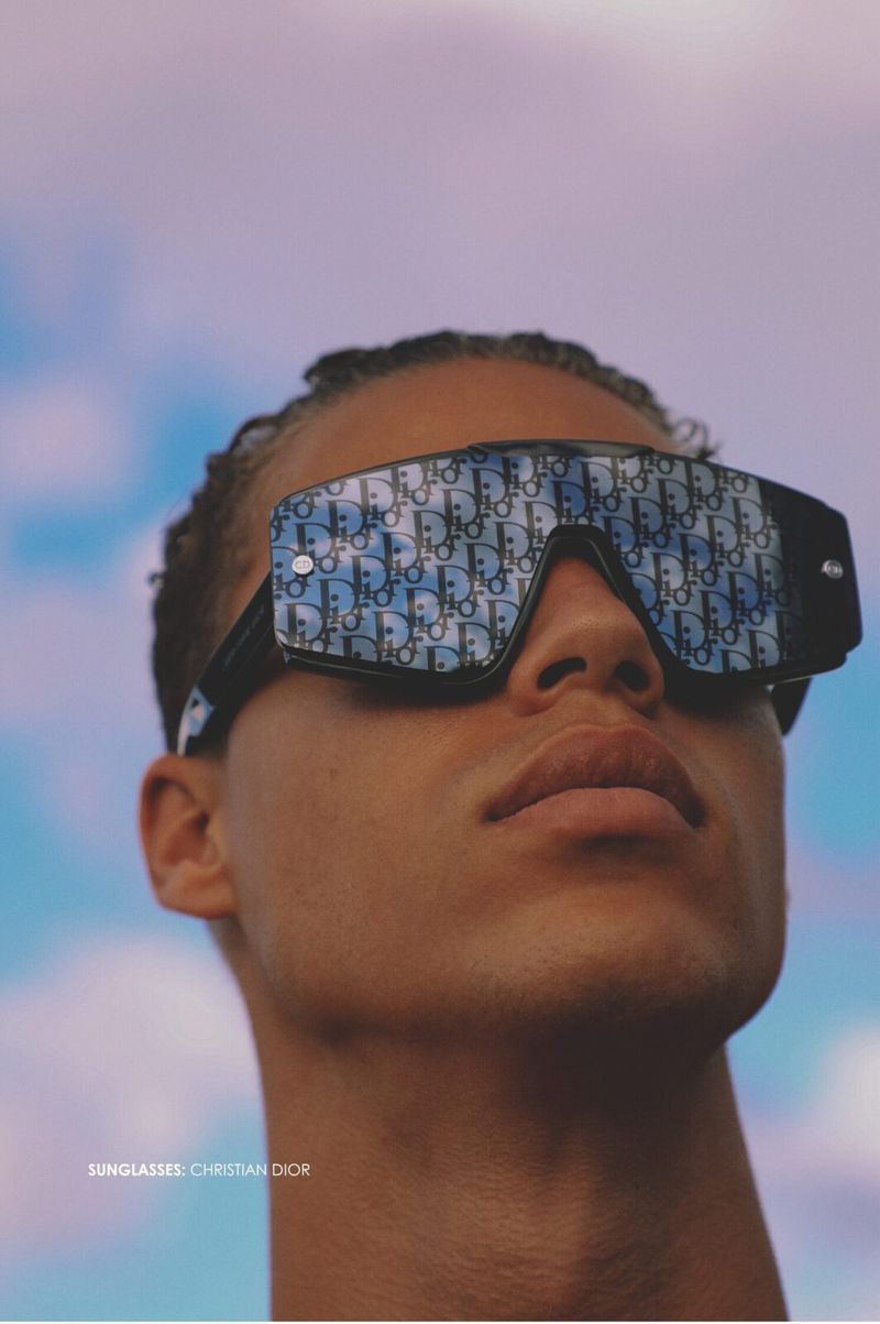Low-angle close-up of a man wearing oversized Christian Dior monogram shield sunglasses against a soft blue sky.