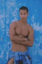 Shirtless man with arms crossed leaning against a weathered blue textured wall with a denim shirt tied at his waist.