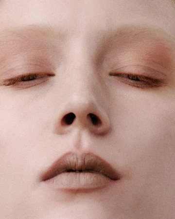Extreme close-up portrait of a woman with closed eyes, bleached eyebrows, and neutral nude makeup filling the frame.