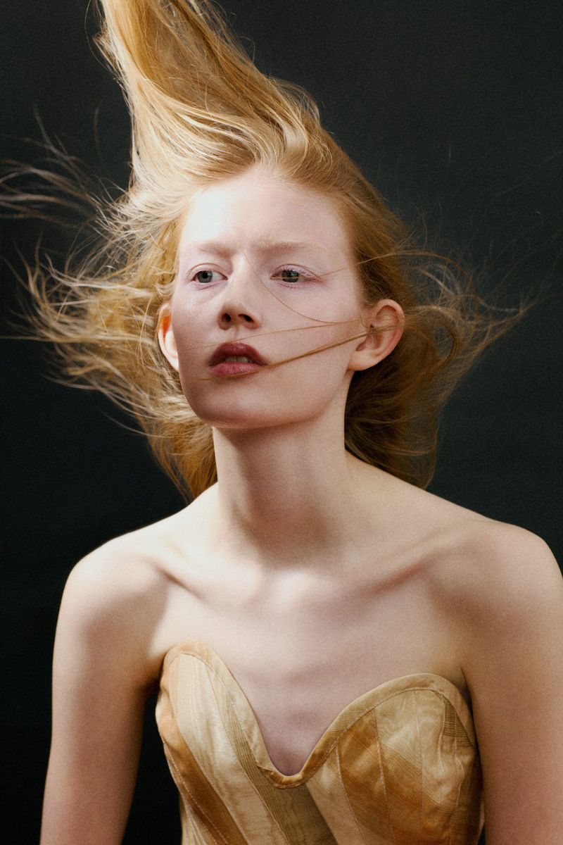 Model with windswept strawberry blonde hair wearing a strapless champagne corset against a black background.