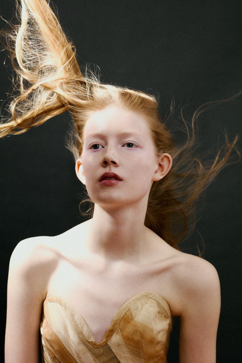 Model with windblown strawberry blonde hair wearing a strapless tan structured top against a black studio background.