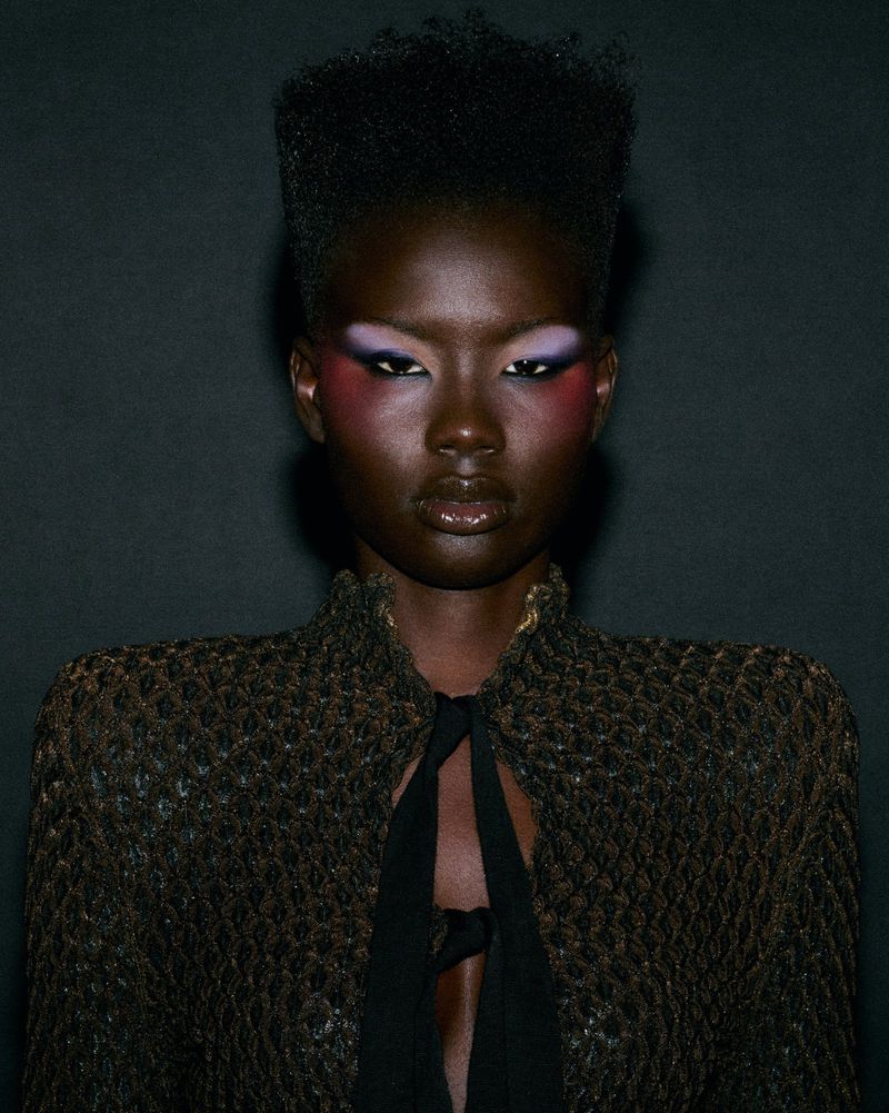 Black woman with high-top fade hair, purple and pink eye makeup, and a textured open jacket against a dark background.