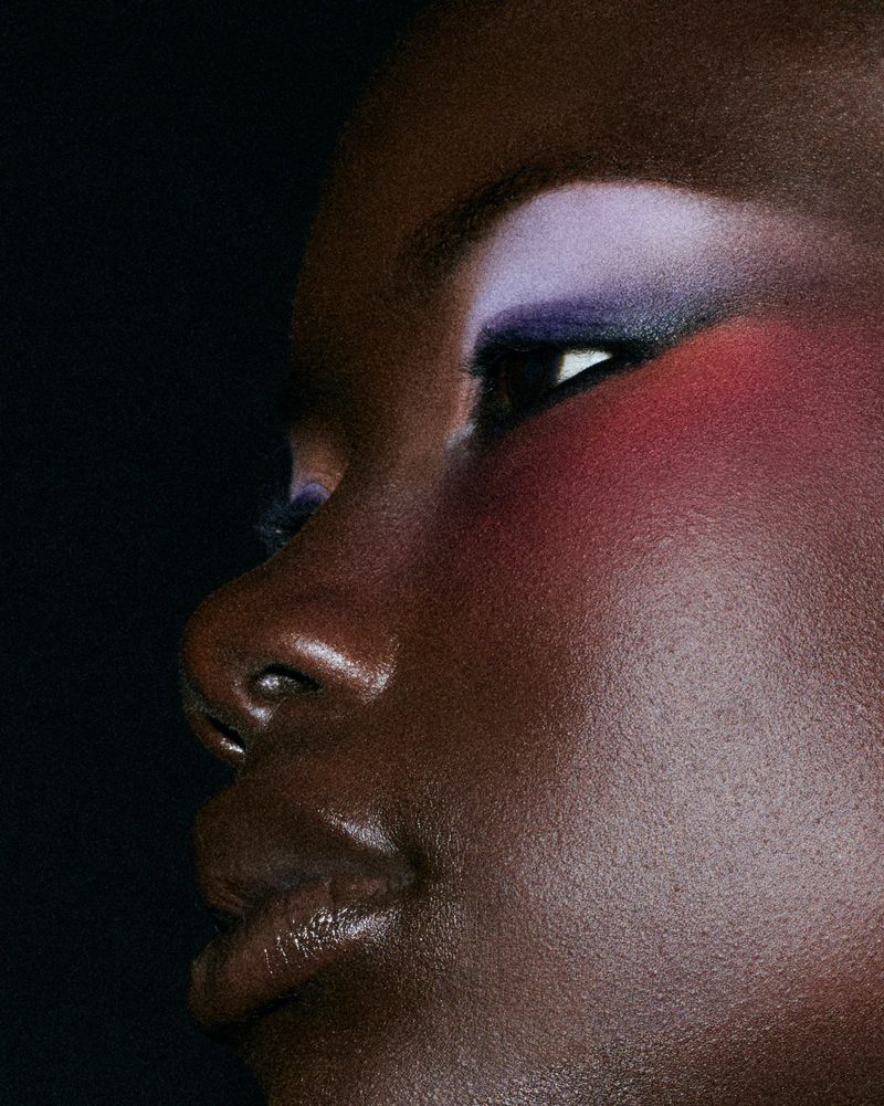 Extreme close up of a Black woman with vibrant lavender eyeshadow and bold pink blush against a dark backdrop.