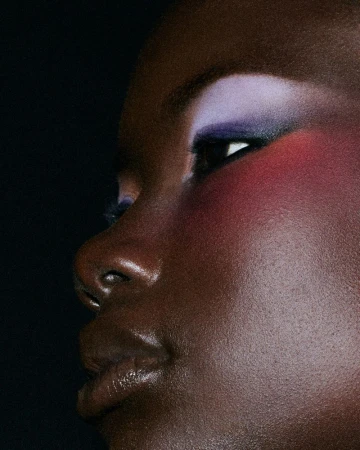 Extreme close up of a Black woman with vibrant lavender eyeshadow and bold pink blush against a dark backdrop.