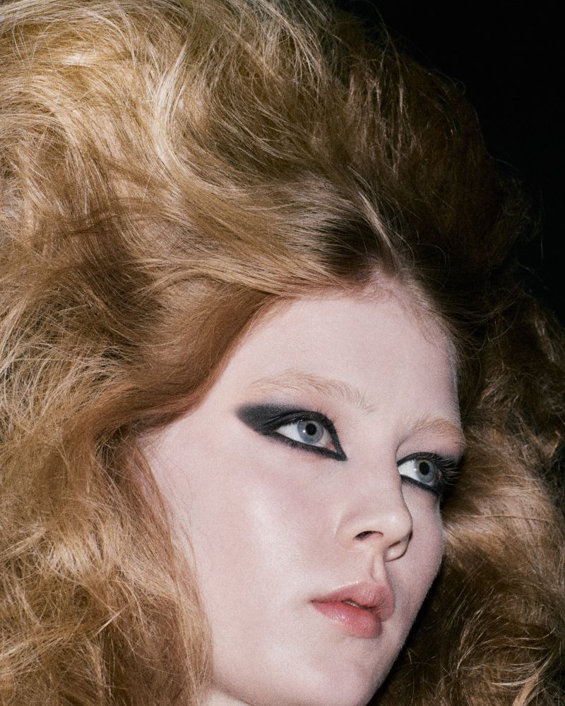 Close-up beauty portrait of a woman with dramatic voluminous teased hair and bold black winged graphic eye makeup against a dark background.