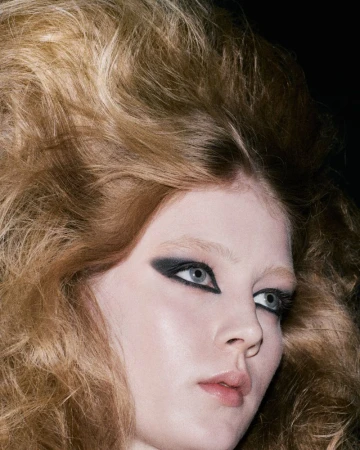 Close-up beauty portrait of a woman with dramatic voluminous teased hair and bold black winged graphic eye makeup against a dark background.