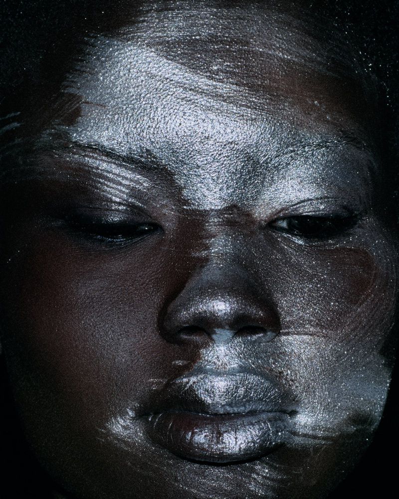 Close-up beauty portrait of a Black model with textured metallic silver makeup brushed across her face against a black background.