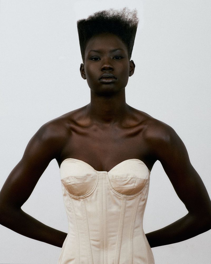 Black woman with a flat-top hairstyle wearing a cream strapless corset top against a plain studio background.