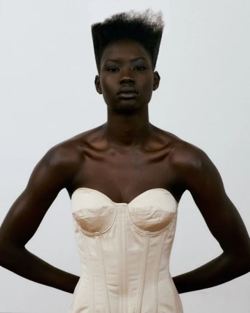 Black woman with a flat-top hairstyle wearing a cream strapless corset top against a plain studio background.