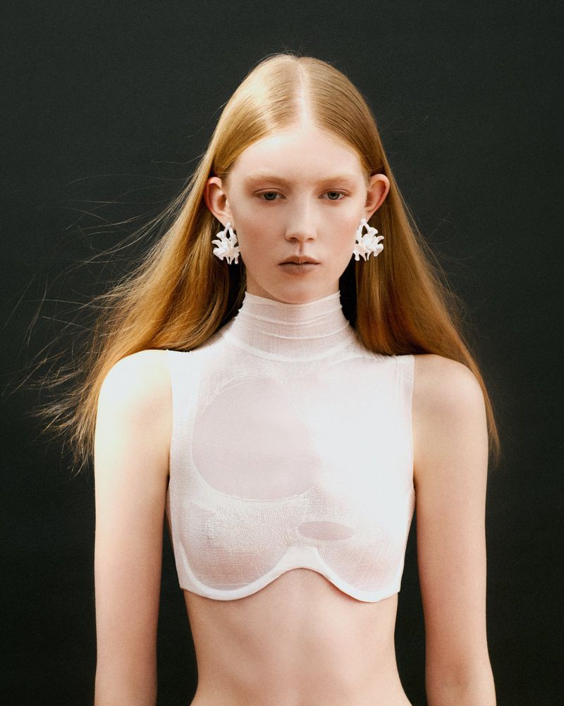 Model with long strawberry blonde hair wearing a sheer white high-neck cropped top and white statement earrings against a black backdrop.