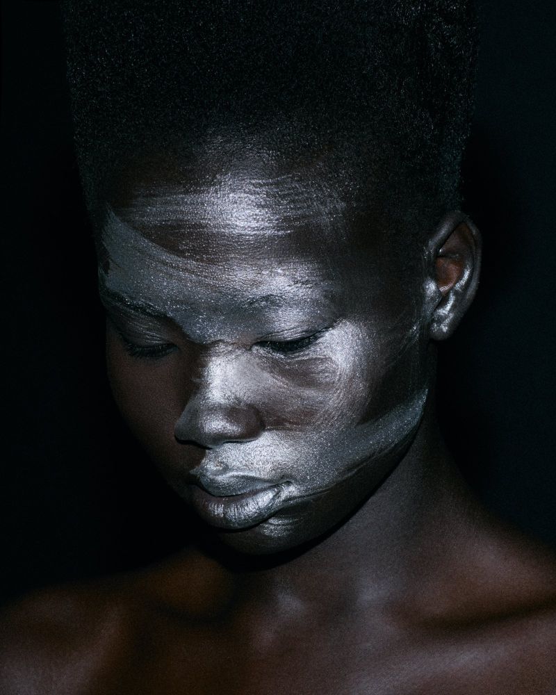 Black model with metallic silver paint brushed across her face against a dark seamless backdrop.