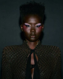 Black woman with high-top fade hair, purple and pink eye makeup, and a textured open jacket against a dark background.