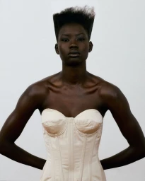 Black woman with a flat-top hairstyle wearing a cream strapless corset top against a plain studio background.