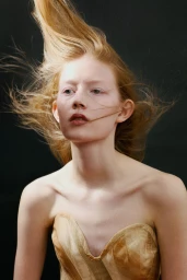 Model with windswept strawberry blonde hair wearing a strapless champagne corset against a black background.