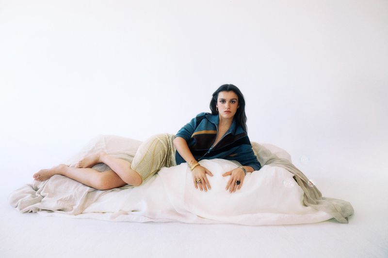 Amaia Romero reclining on draped fabric wearing a teal zip jacket and shimmering beige skirt against a white studio background.