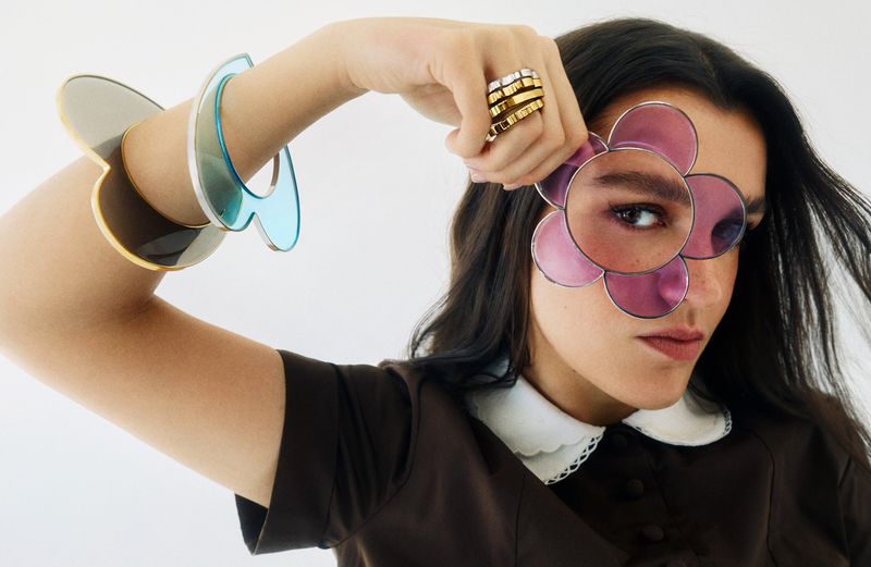 Amaia Romero looking through a translucent pink flower-shaped glass prop while wearing oversized sculpted bangles and a collared dress.