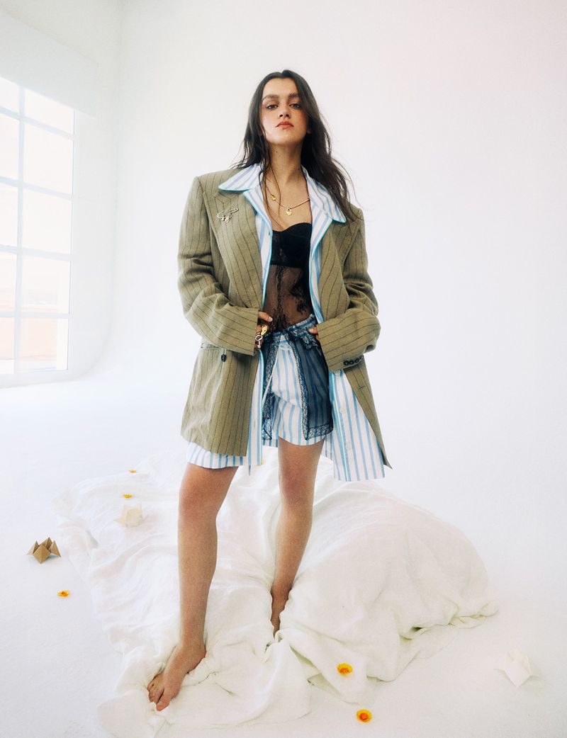 Amaia Romero standing barefoot wearing an oversized pinstriped blazer, striped shirt, and black lace top against a white studio background.