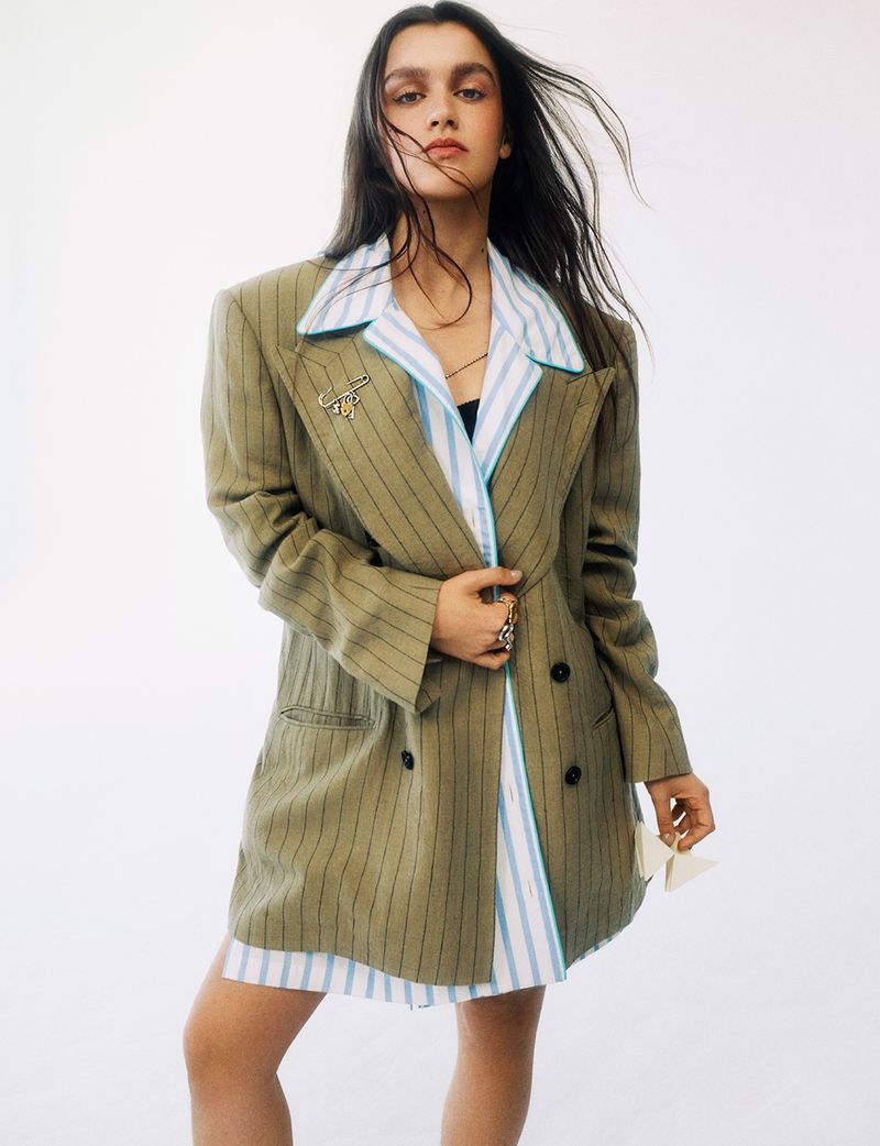 Amaia Romero posing in an oversized olive pinstriped blazer over a striped collared shirt against a white backdrop.