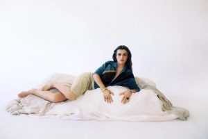 Amaia Romero reclining on draped fabric wearing a teal zip jacket and shimmering beige skirt against a white studio background.