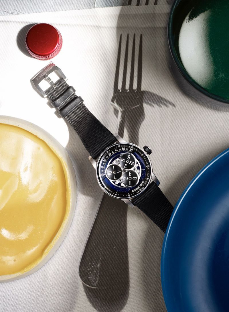 Luxury wristwatch with a black strap placed over a dark fork shadow alongside colorful dishes on a linen surface.