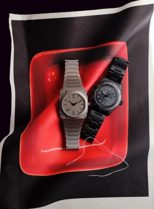 Two luxury wristwatches, one titanium and one matte black, arranged on a red and black printed textile.