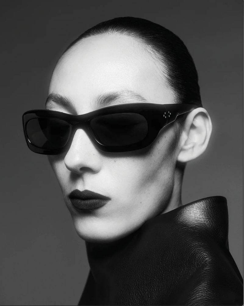 Black and white portrait of a woman wearing oversized black sunglasses and a black leather top against a neutral grey backdrop.