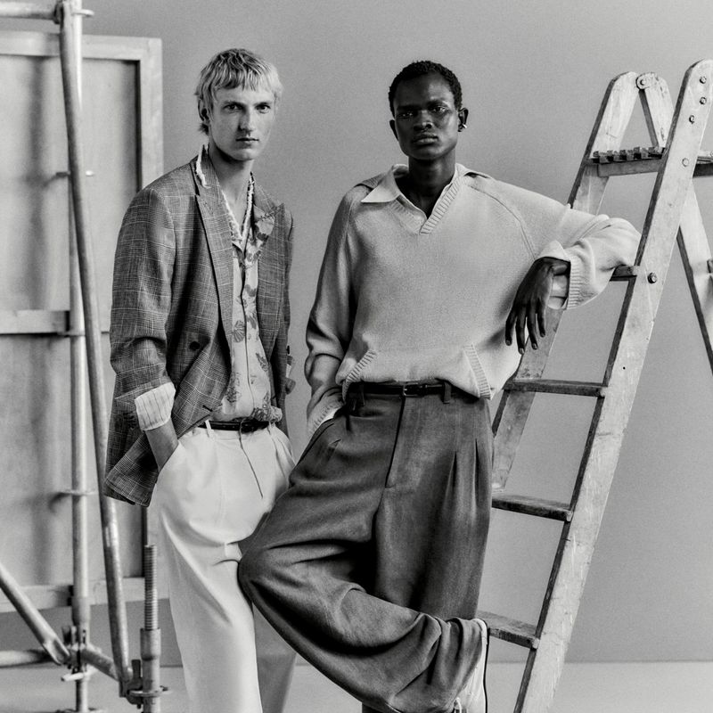 Two male models in tailored menswear posing beside studio scaffolding and a wooden ladder in a monochrome portrait.