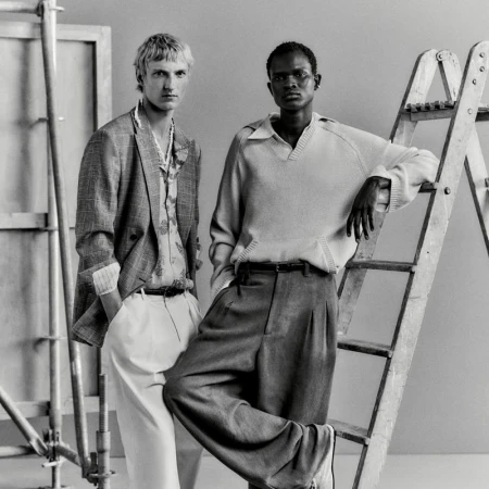 Two male models in tailored menswear posing beside studio scaffolding and a wooden ladder in a monochrome portrait.