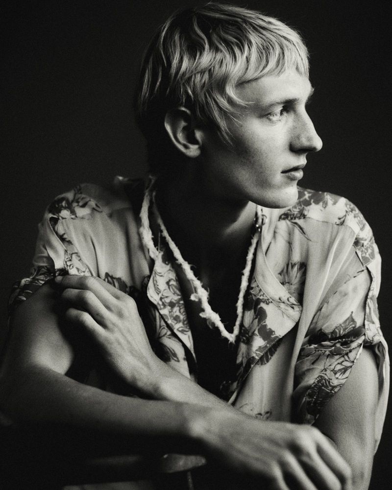 Monochrome portrait of a young man looking to the side, wearing a short-sleeve floral printed shirt against a dark background.
