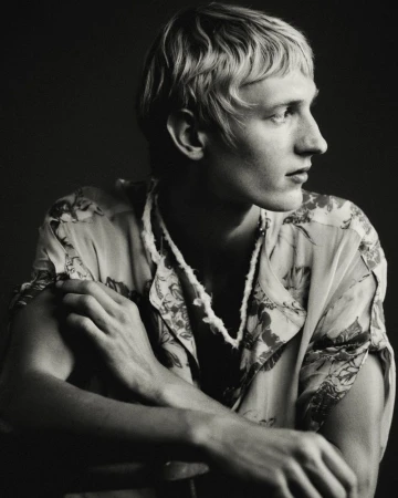 Monochrome portrait of a young man looking to the side, wearing a short-sleeve floral printed shirt against a dark background.
