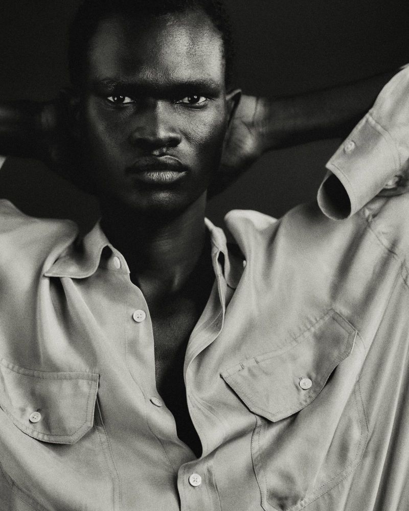 Black and white portrait of model Chubath Kutien wearing an unbuttoned collared shirt with hands behind his head.