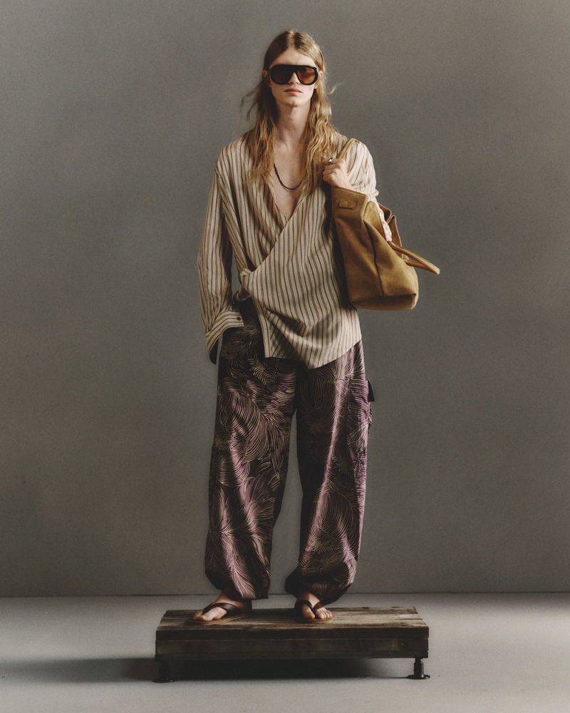 Model wearing dark shield sunglasses, a striped wrap shirt, printed trousers, and holding a tan tote bag on a low wooden platform.