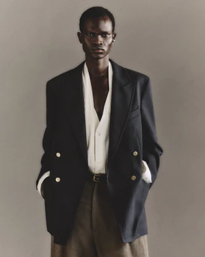 Model Chubath Kutien wearing an oversized navy double-breasted blazer, unbuttoned white shirt, and brown trousers against a grey backdrop.