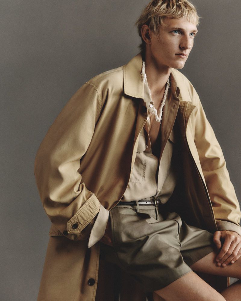 Blond male model wearing an open beige trench coat, linen shirt, and tailored shorts against a grey studio backdrop.