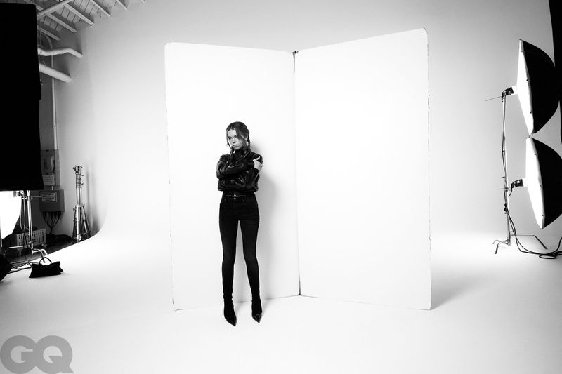 Milly Alcock standing in an all-black leather outfit in front of white studio flats with lighting equipment visible around her.