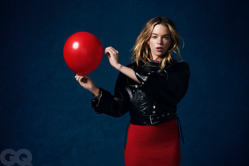 Milly Alcock holding a red balloon wearing a black leather jacket and red skirt against a dark blue background.