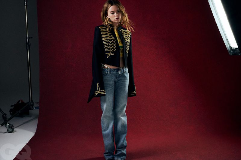 Milly Alcock stands against a burgundy studio backdrop wearing a black embroidered military jacket and blue denim jeans.