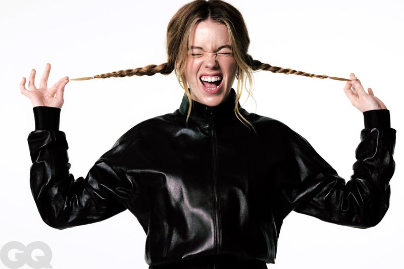 Milly Alcock pulling her braided hair outwards while laughing in a black leather jacket against a white studio background.