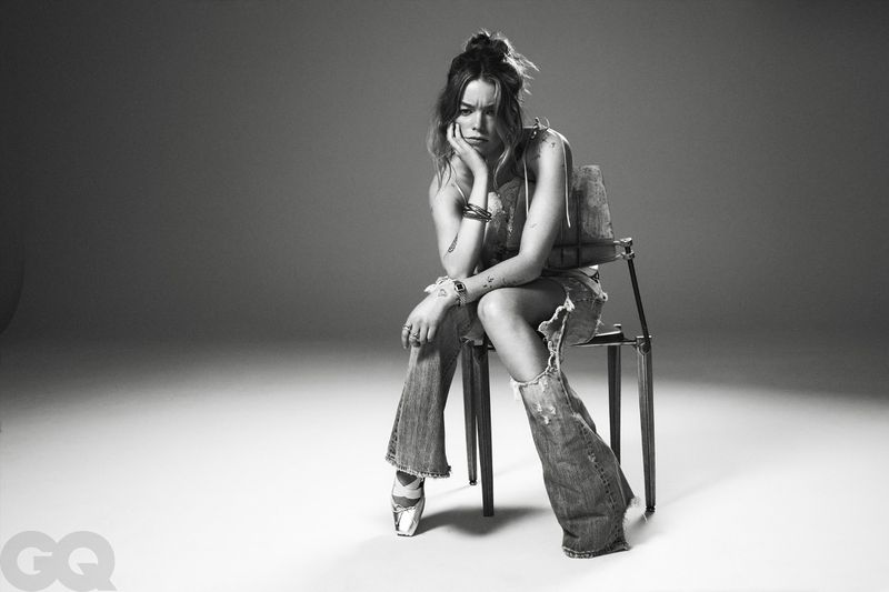 Milly Alcock resting her chin on her hand while seated on a wooden chair wearing distressed flared jeans against a studio backdrop.