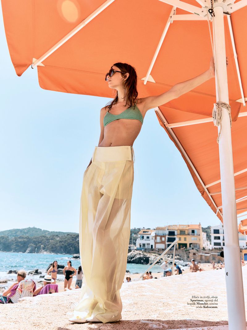 Model Tani Birkin stands under an orange beach umbrella wearing a green bikini top and sheer pale yellow trousers on a sunny beach.