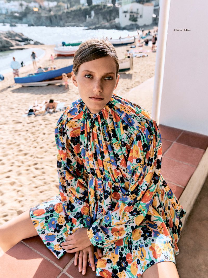 Model wearing a colourful floral dress seated outdoors on a red tiled ledge overlooking a beach and sea