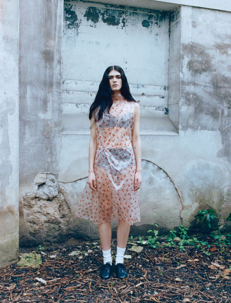 Model Angelina Sandoval poses in a sheer printed sleeveless dress and dark undergarments against a weathered concrete wall.