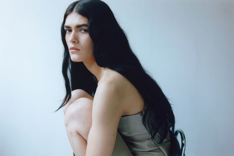 Model Angelina Sandoval crouching in a grey dress with long flowing black hair against a neutral studio backdrop.
