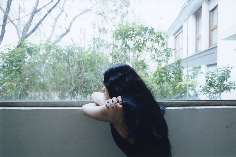 A woman with long black hair rests her arms on a ledge while looking out a large glass window toward lush green trees.