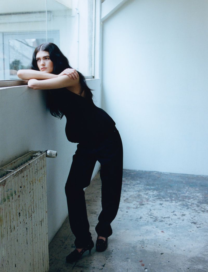 Model with long dark hair leaning on a window sill wearing a black top and trousers in a sparse interior space
