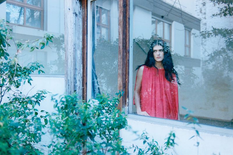 Model Angelina Sandoval in a red dress looking out from behind a reflective glass window framed by green foliage.
