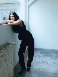 Model with long dark hair leaning on a window sill wearing a black top and trousers in a sparse interior space