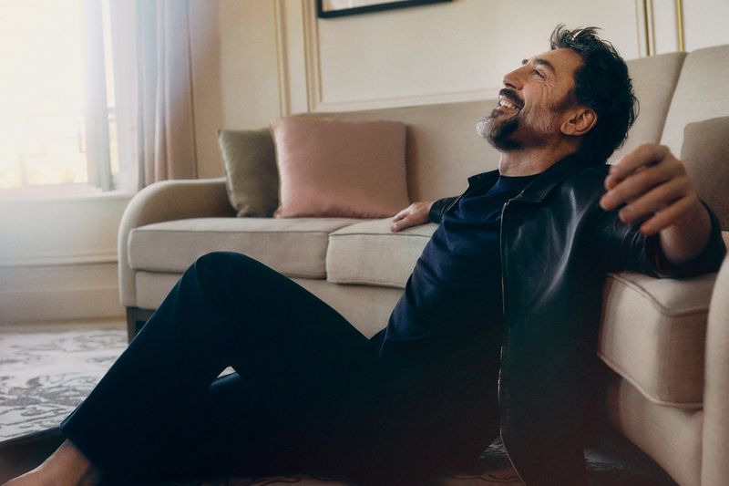 Actor Javier Bardem laughing while sitting on the floor leaning against a beige sofa in a sunlit room.